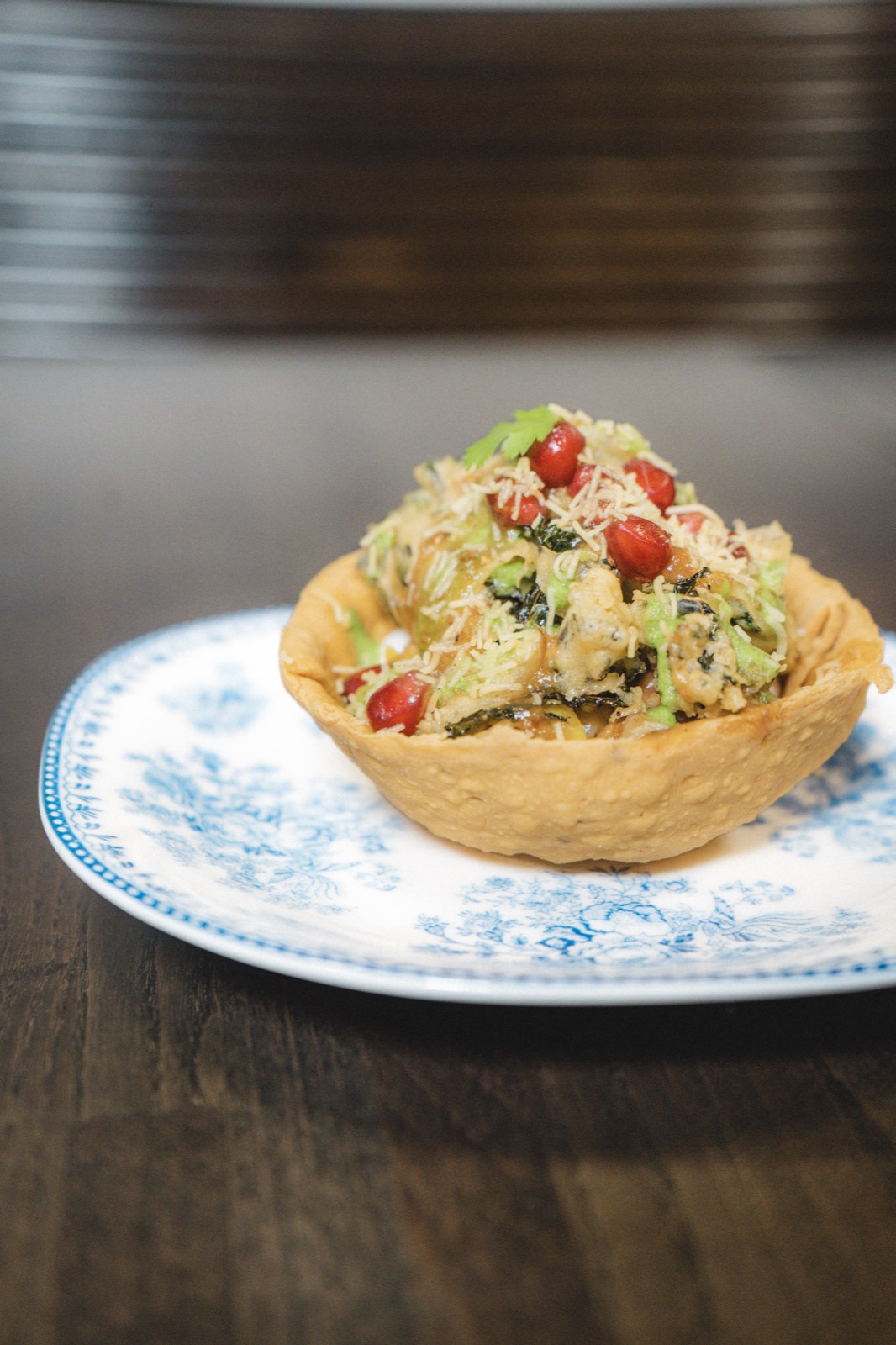 A fried pastry cup filled with greens, crisp noodles and pomegranate seeds on a blue and white plate at Chhauka